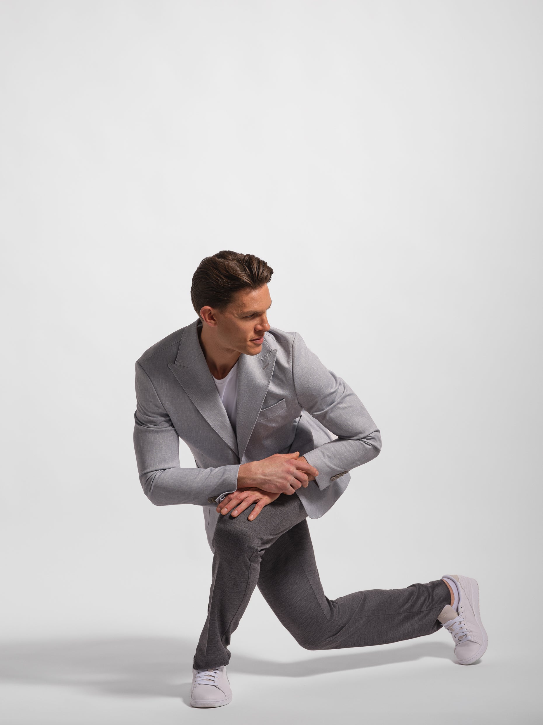 Man in a gray blazer showing stretch and white sneakers posing on a light gray background