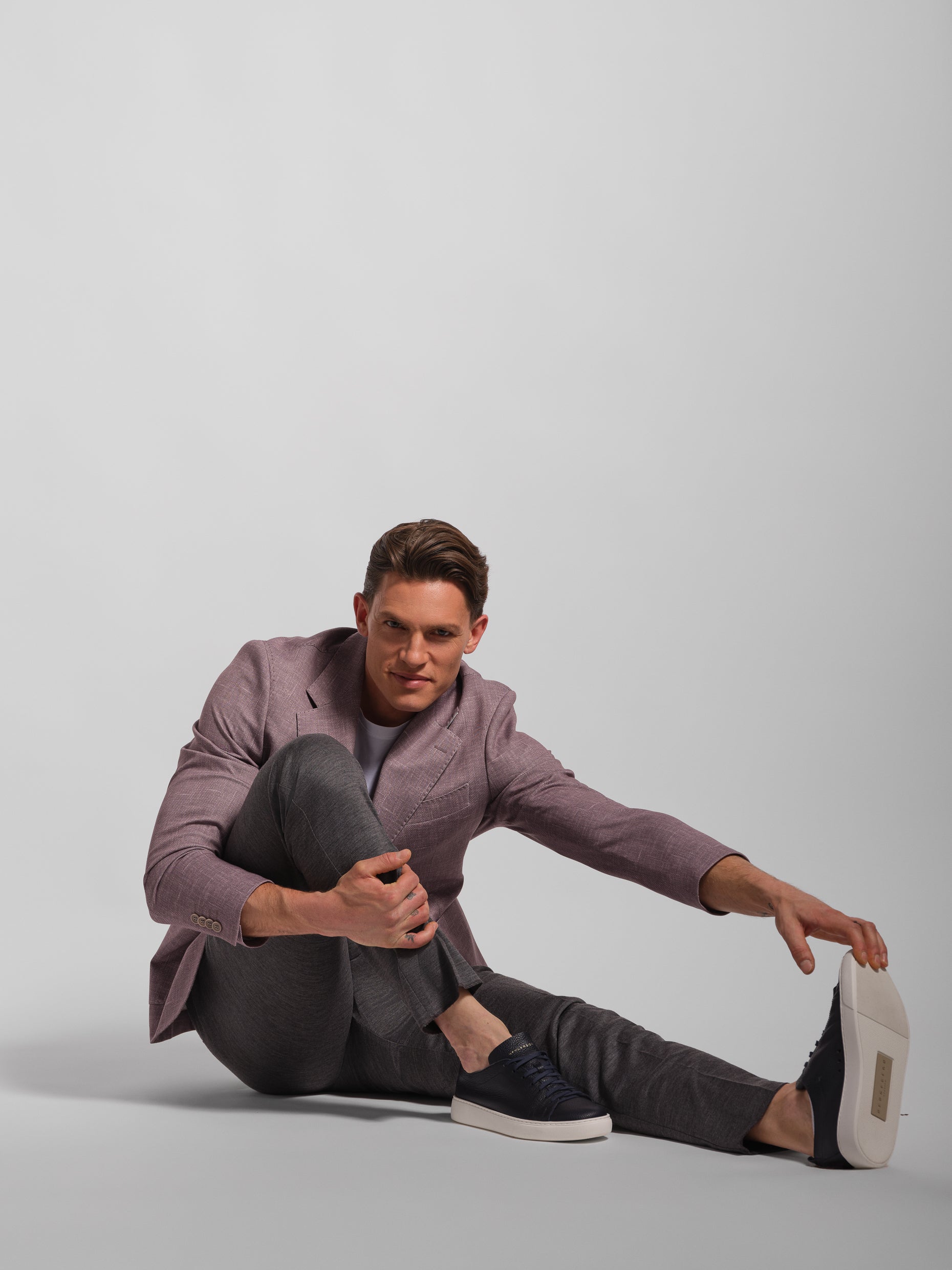 Man sitting on the floor wearing a mauve stretch and comfortable blazer jacket and gray pants against a plain background