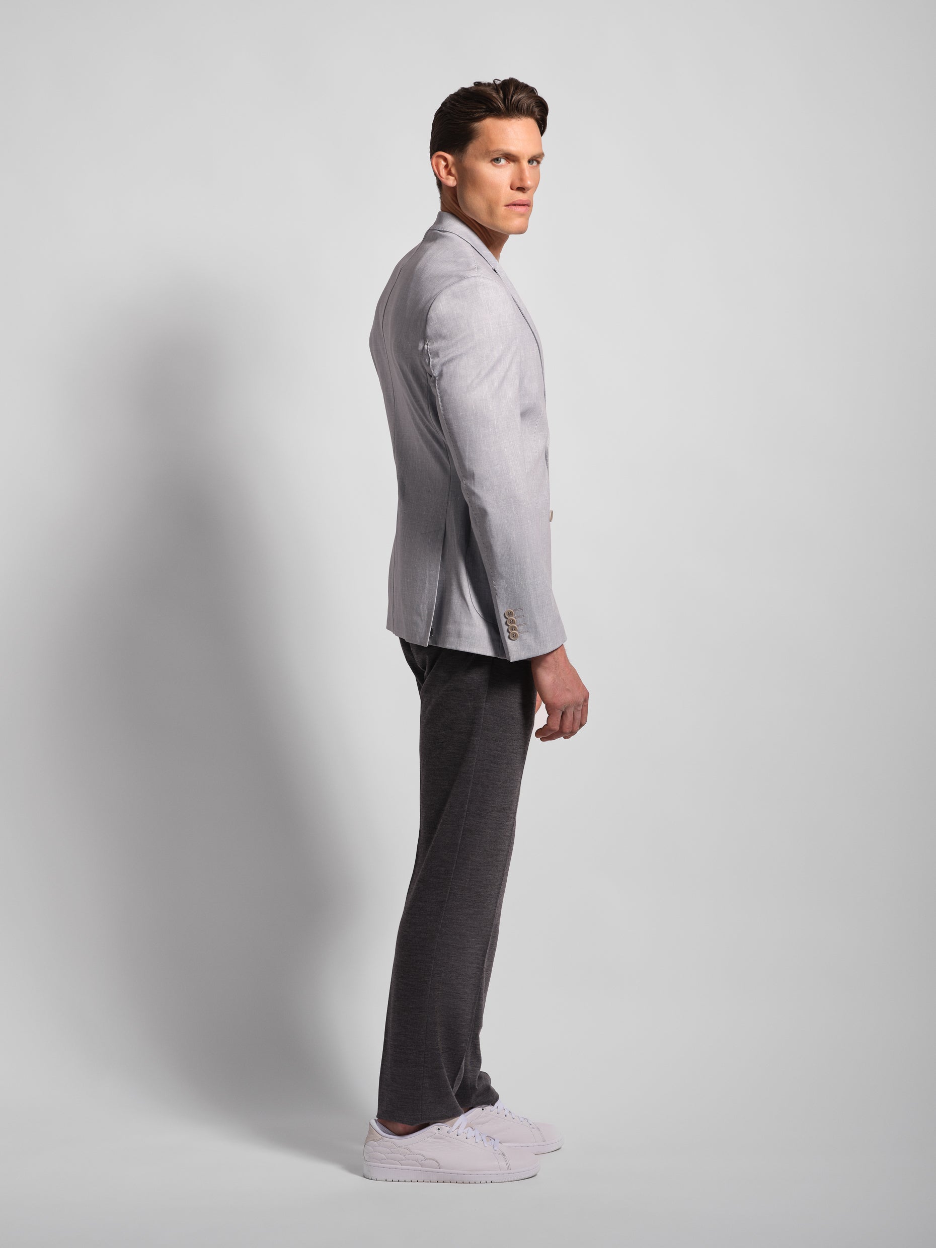 Man wearing a light gray stretch and comfortable jacket blazer by profile side and dark gray pants on a plain background