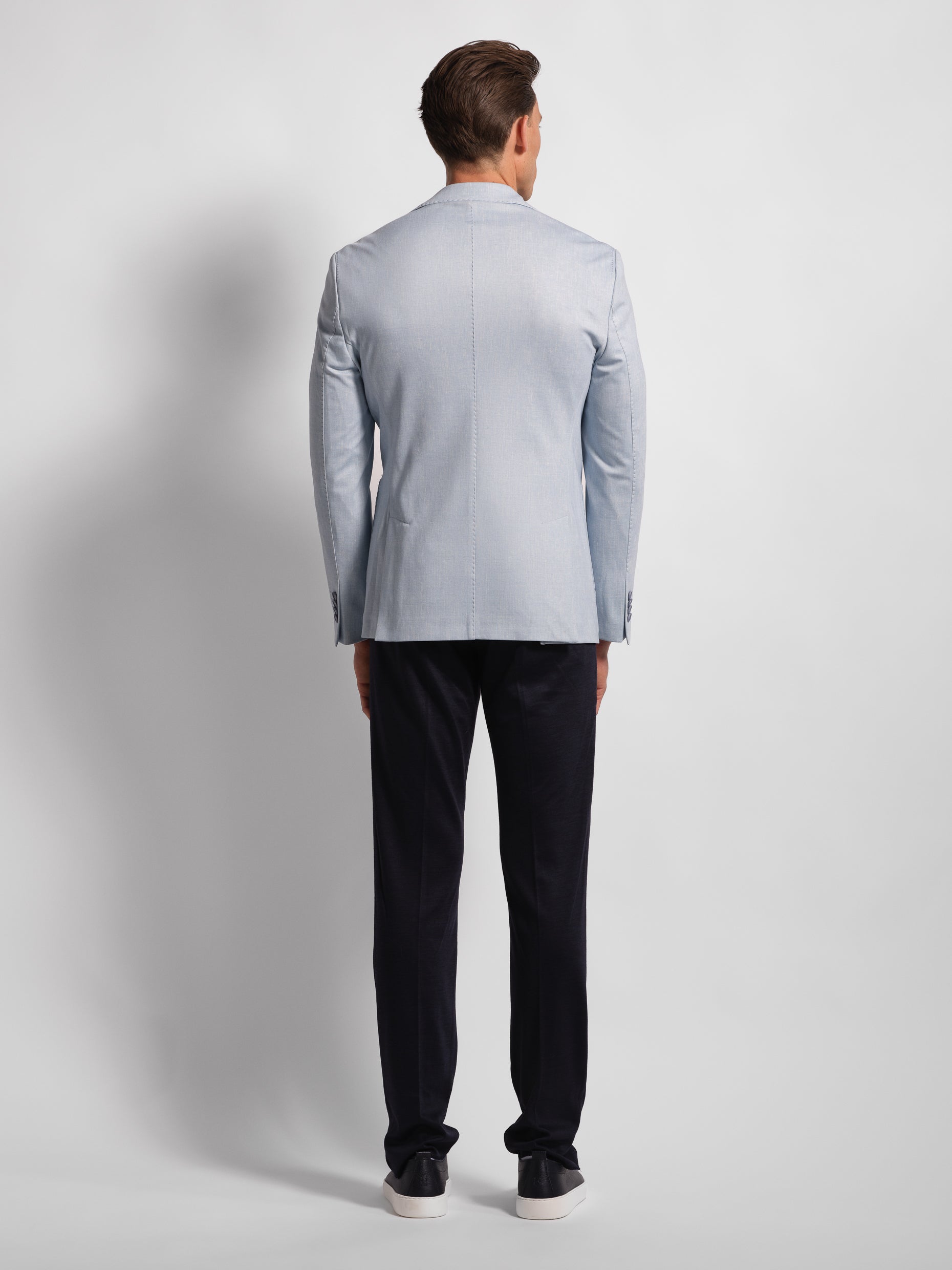 Man wearing a light blue blazer and navy pants on a plain background. showing his backside