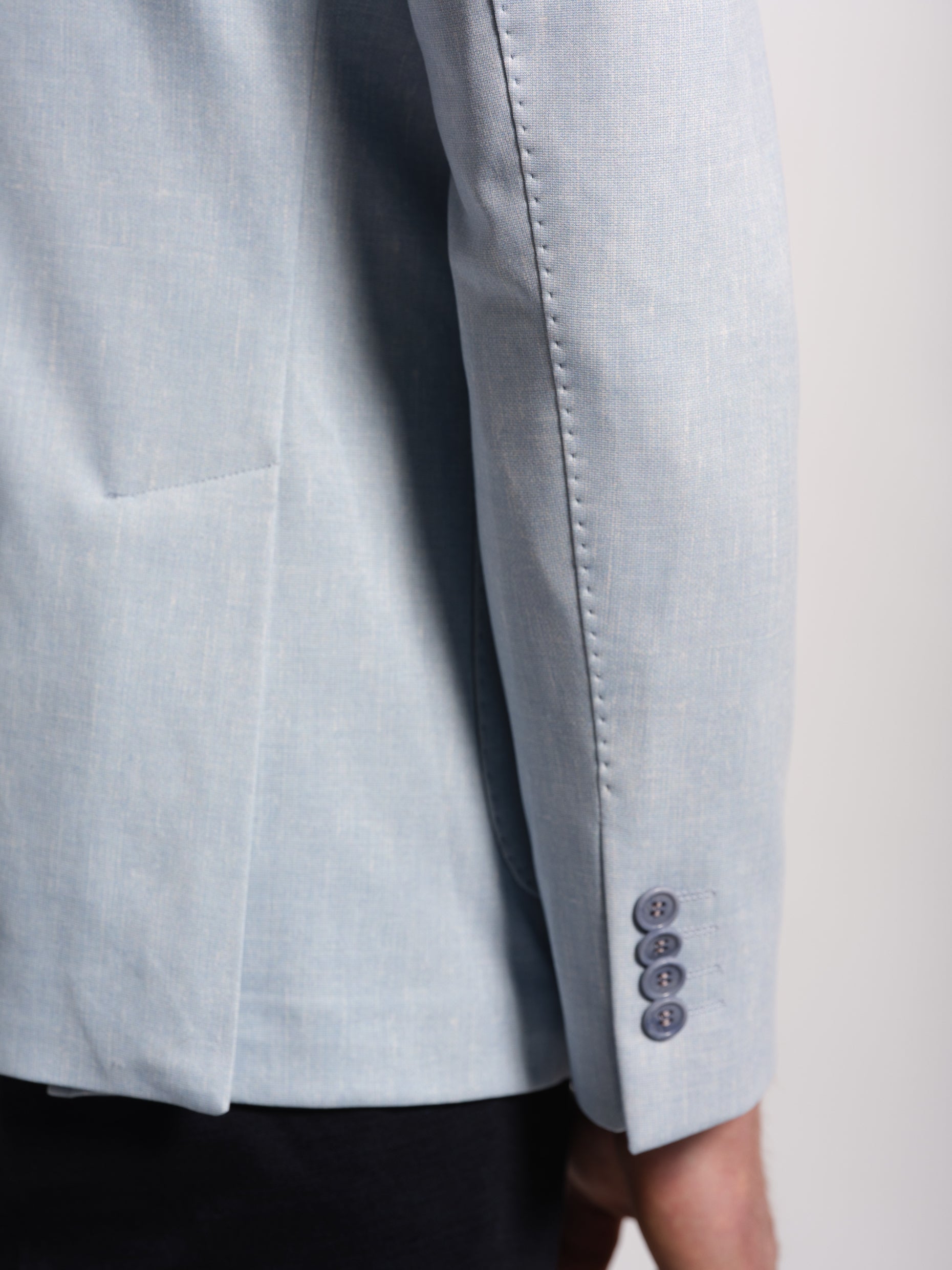 Close-up of a light blue suit jacket with button details on the sleeve , top stiches, a neutral background