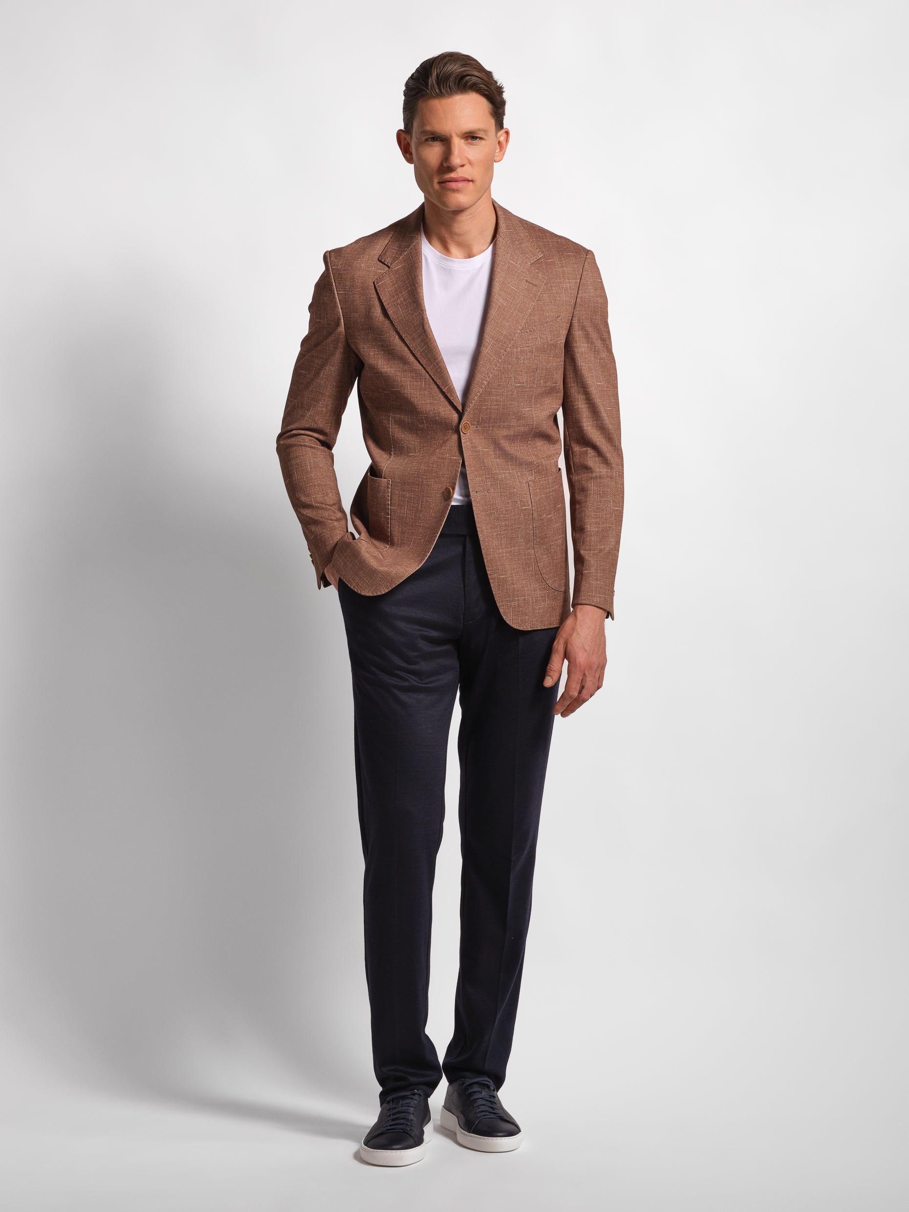 Man in a stretchy, comfortable jersey brown blazer over a white t-shirt with navy pants hand in pocket 